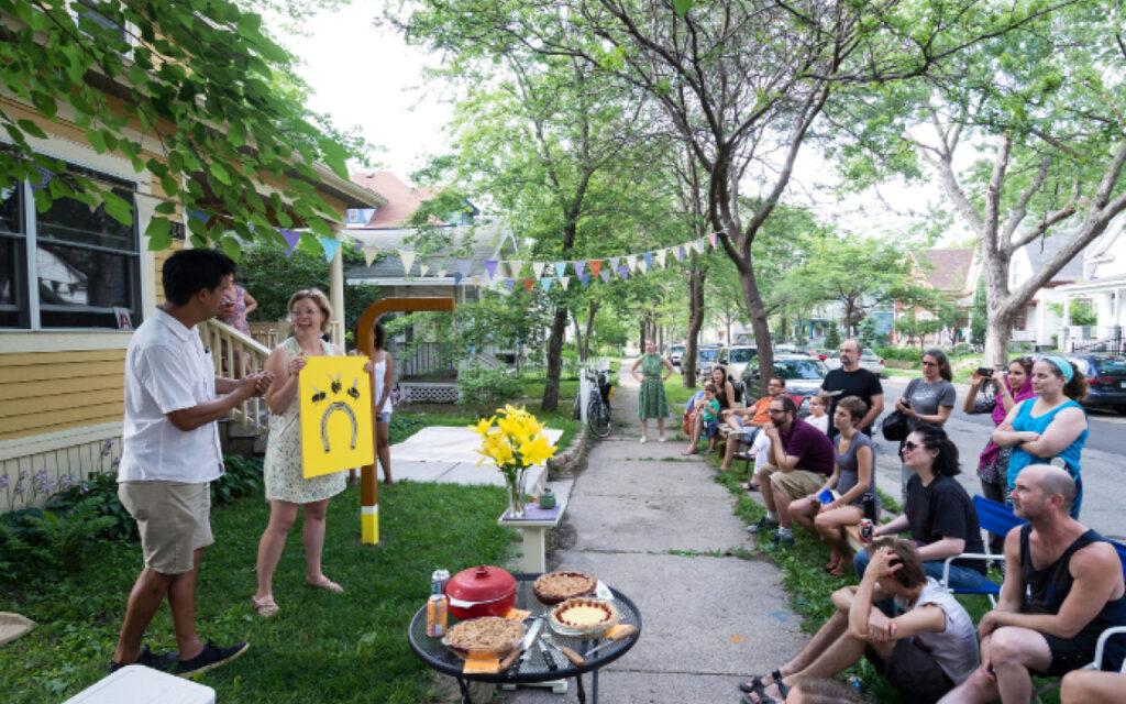 Two people stand on the front lawn of a yellow house on a tree-lined residential street, holding up a bright yellow sign with a drawing on it. A seated audience of about a dozen people watches from lawn chairs and steps. A table with pies, flowers, and drinks is in the foreground. Colorful pennant banners are strung above.