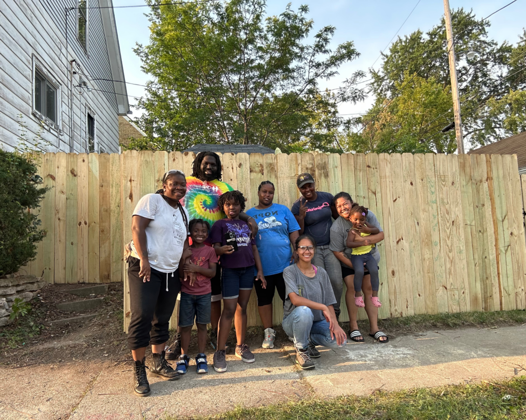 A group of people posing in front of a freshly-built wooden privacy fence.