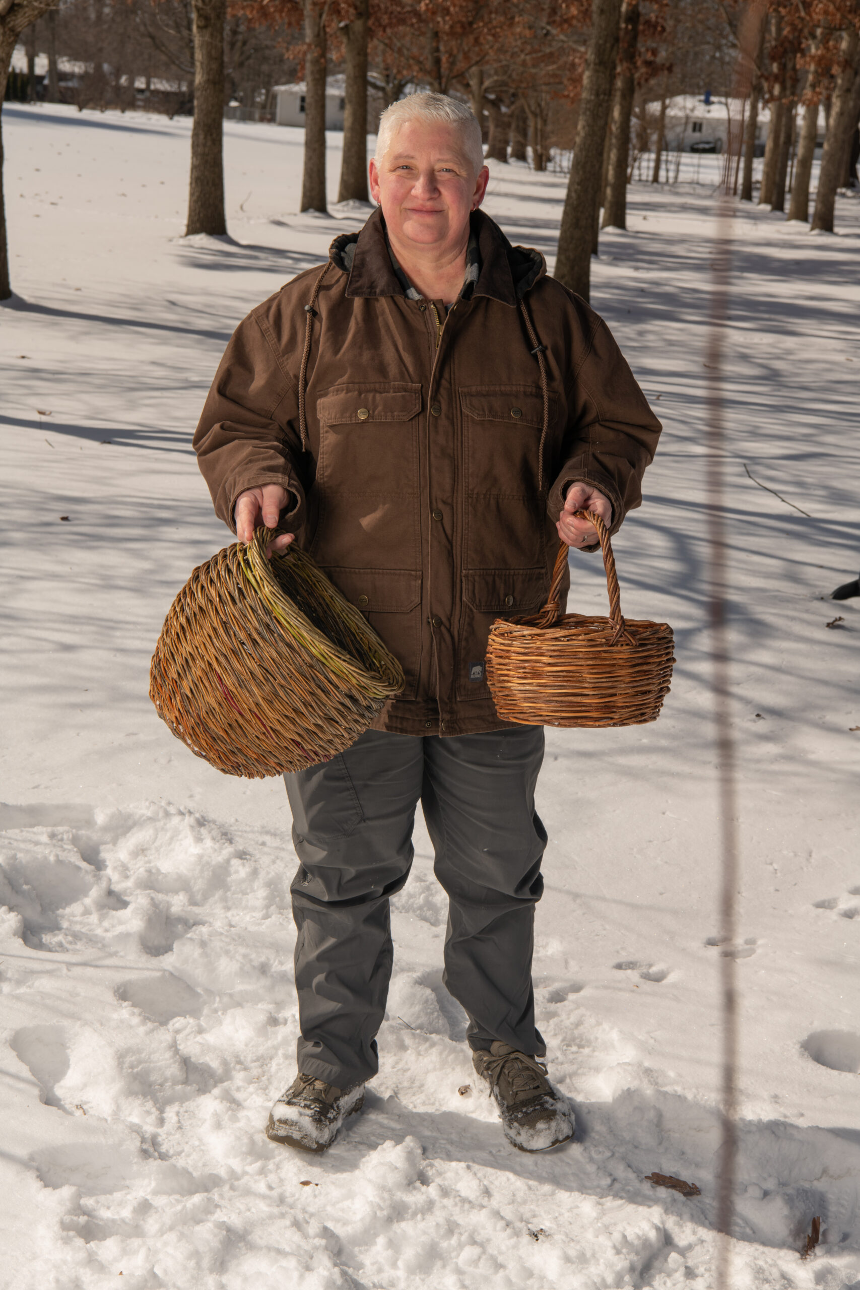 A person with light skin and short white hair holds two baskets and stands in the snow.