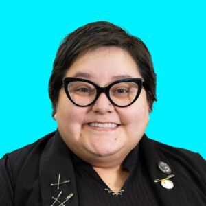 Headshot of a smiling woman w light skin and a black pixie haircut, wearing a black shirt and vest w safety pin embellishments.