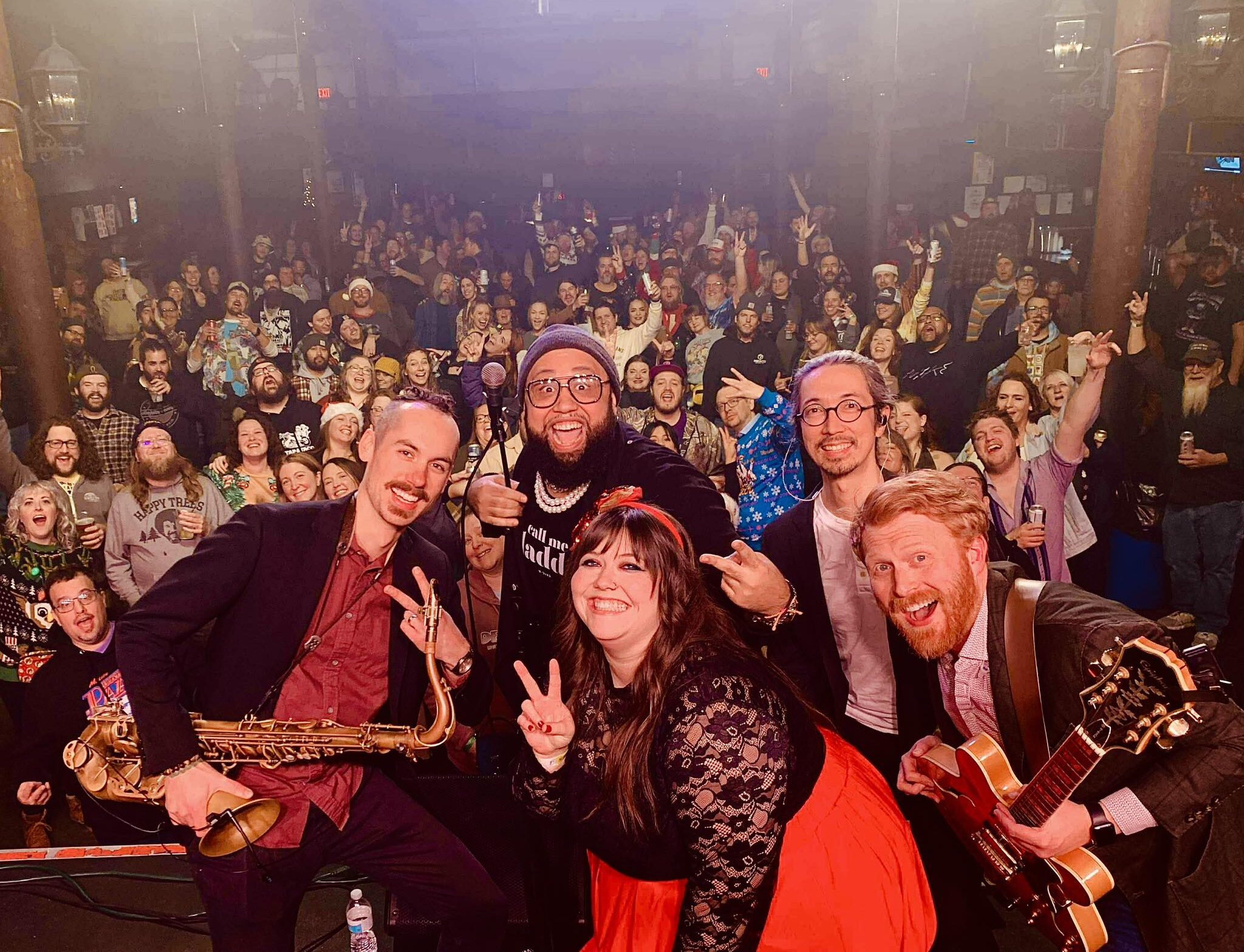 A five-member band smiling and flashing peace signs in front of a live audience.