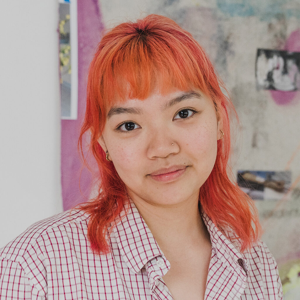 Headshot of a smiling person with light skin tone and shoulder-length orange hair, with gold nose piercing and wearing a checkered pink shirt.