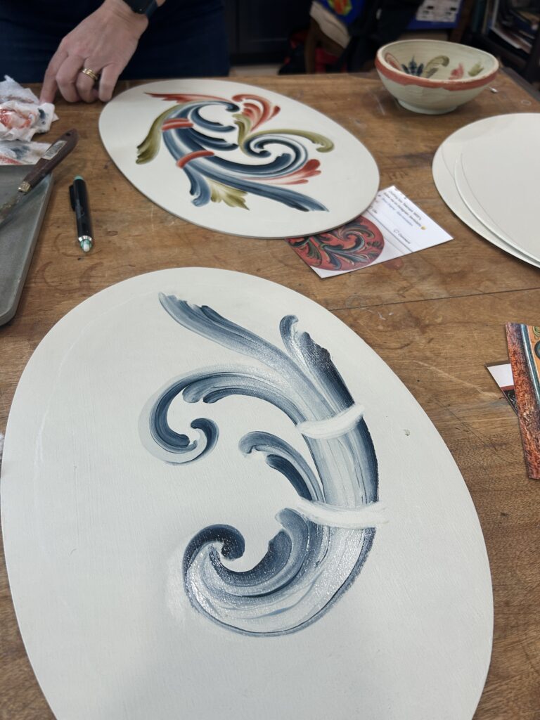 Two pieces of unfired pottery pieces resting on a table as artists paint on them.