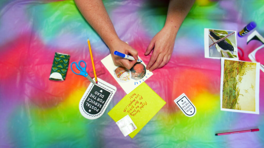 A pair of light-skinned hands drawing on a collage next to scattered art supplies on a colorful table.