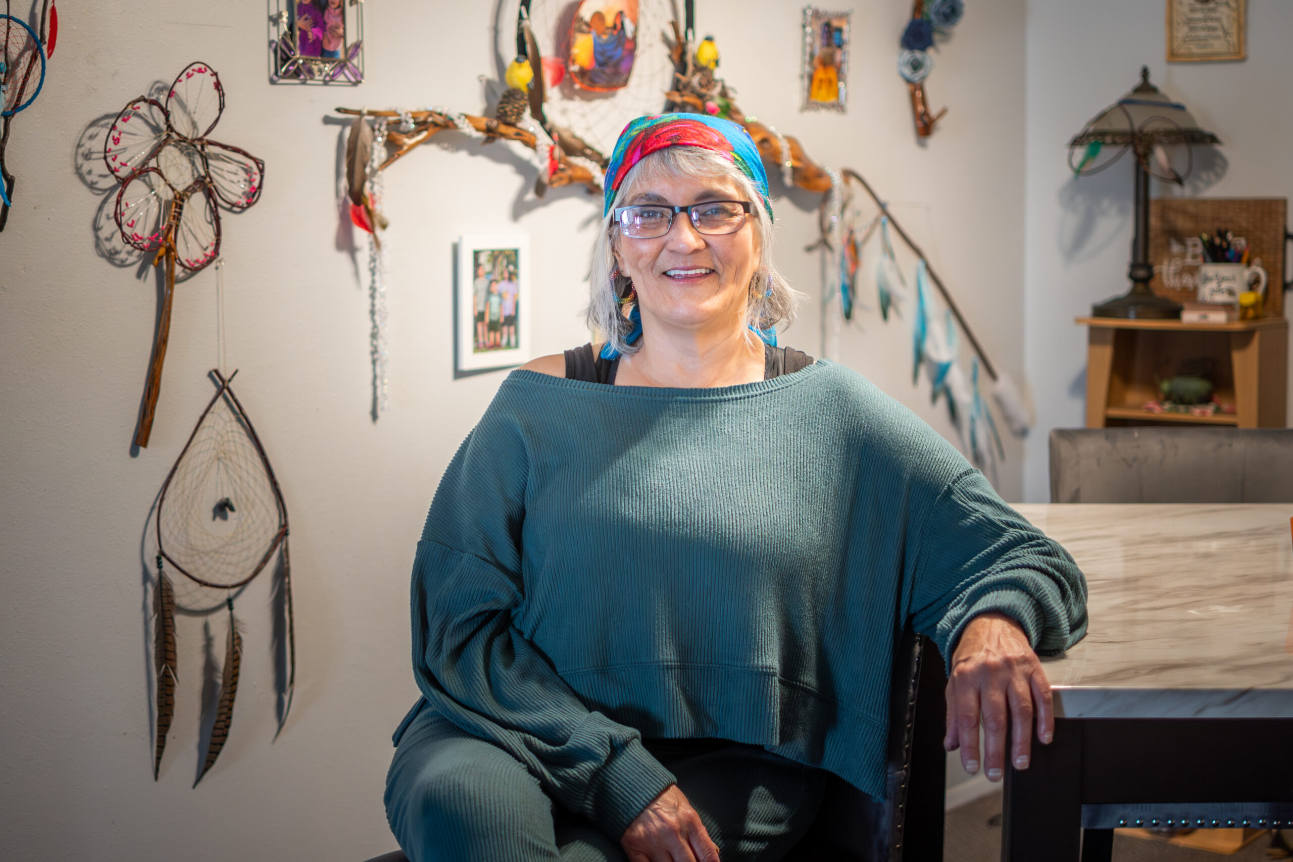 A person of ligh skin tone with a pattern headscarf and glasses smiling and posing for a photo in front of a wall with a variety of dreamcatchers