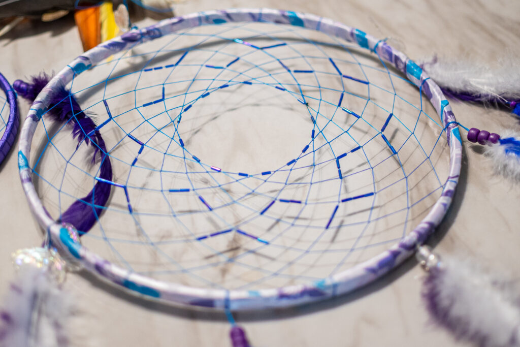 A blue and purple dreamcatcher made with string, beads, paper, and feathers.