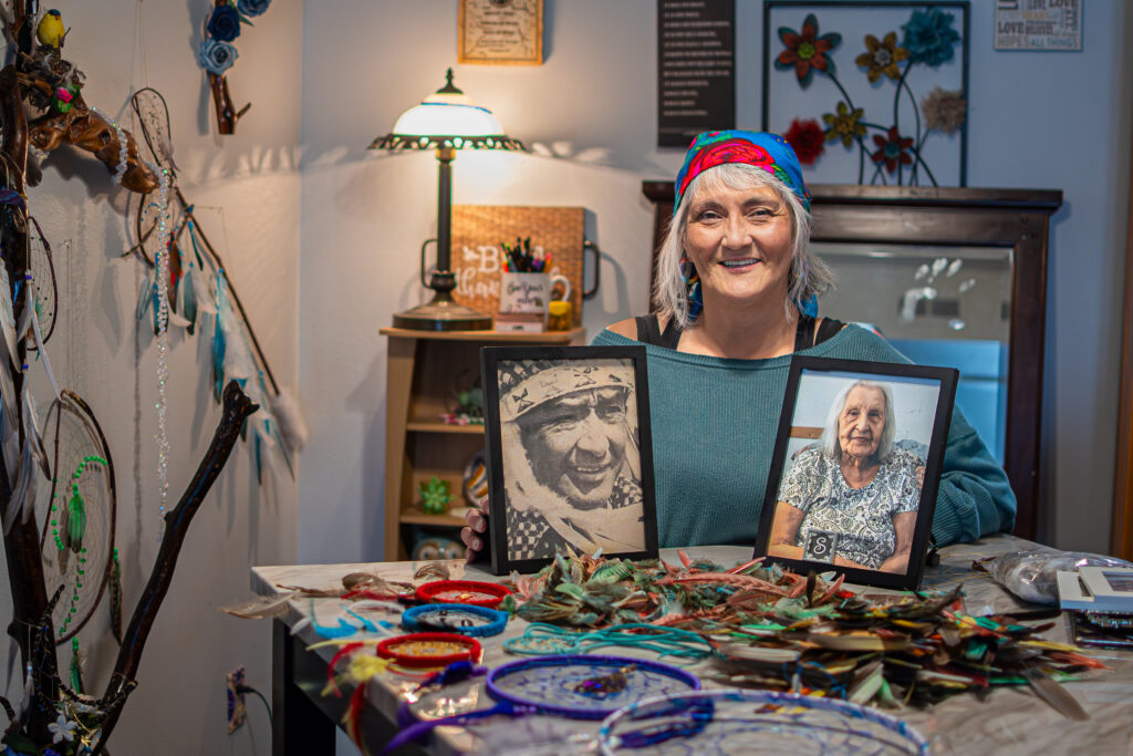 A person of light skin tone sitting at a table with various finished and in-progress dreamcatchers and two framed photos of elders.