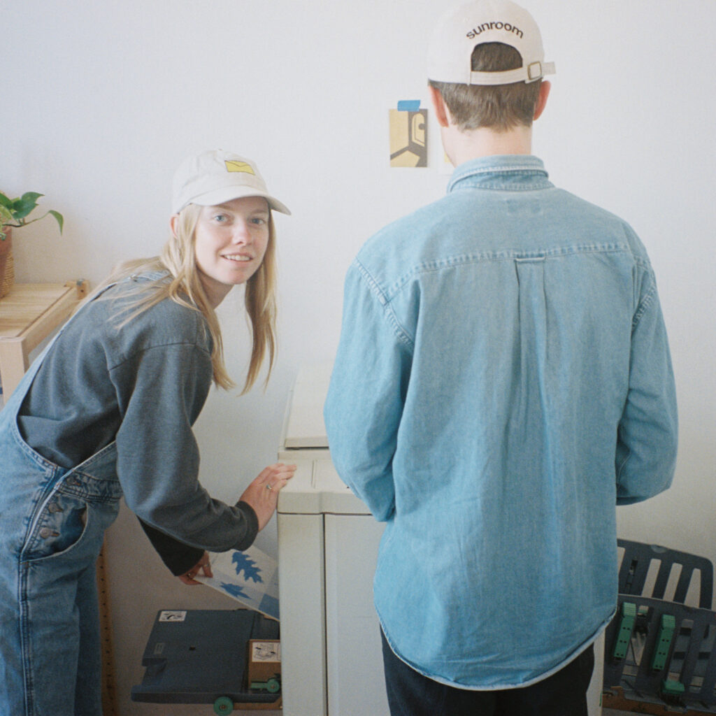 Two people with light skin standing at a risograph printer, one with his back to the camera.