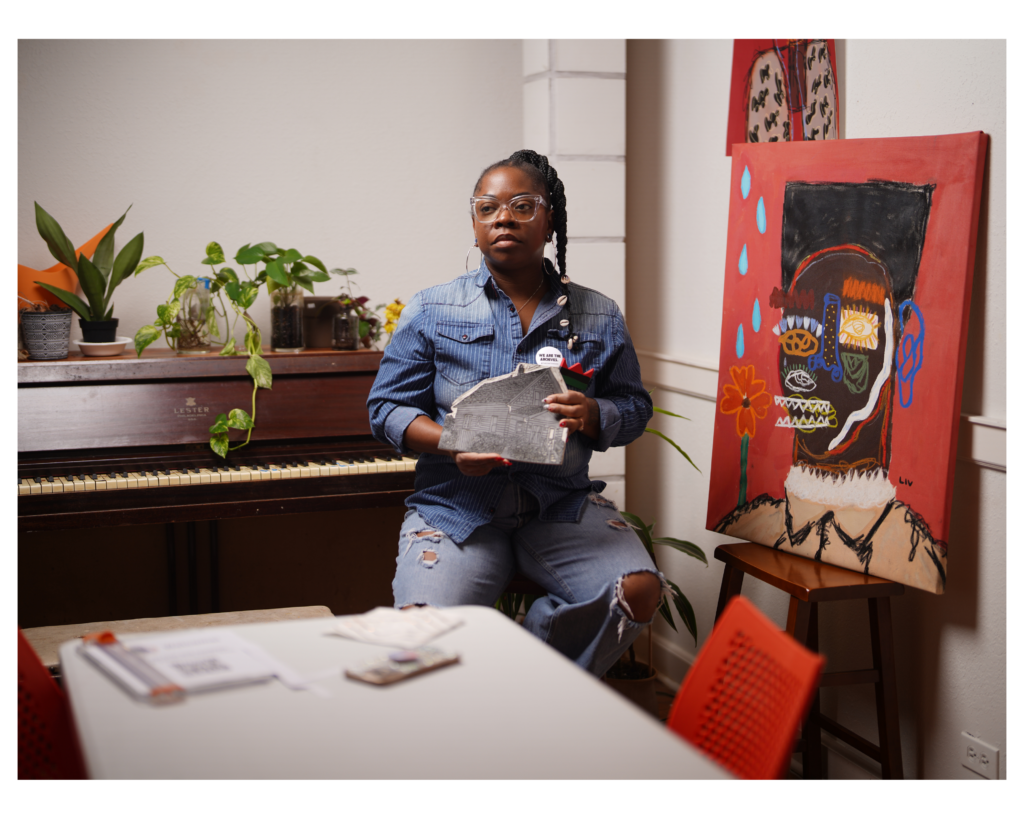 A woman with dark skin tone posing next to a large canvas painting indoors.