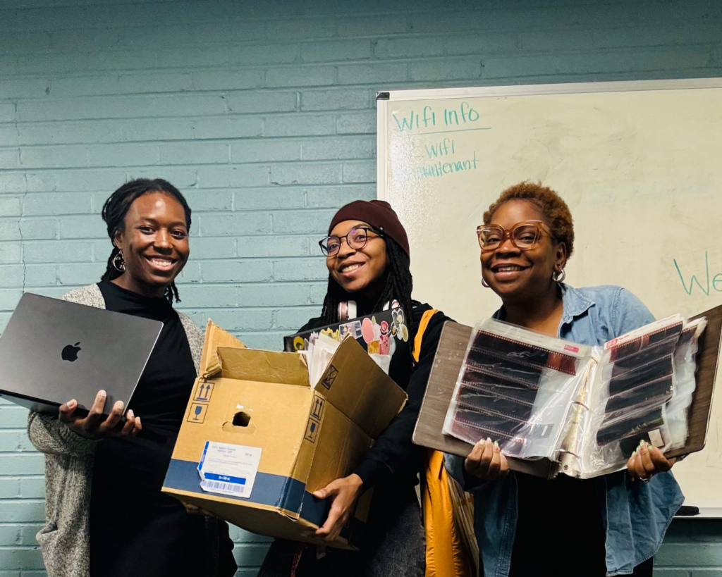 Three women with dark skin smiling and holding photos.