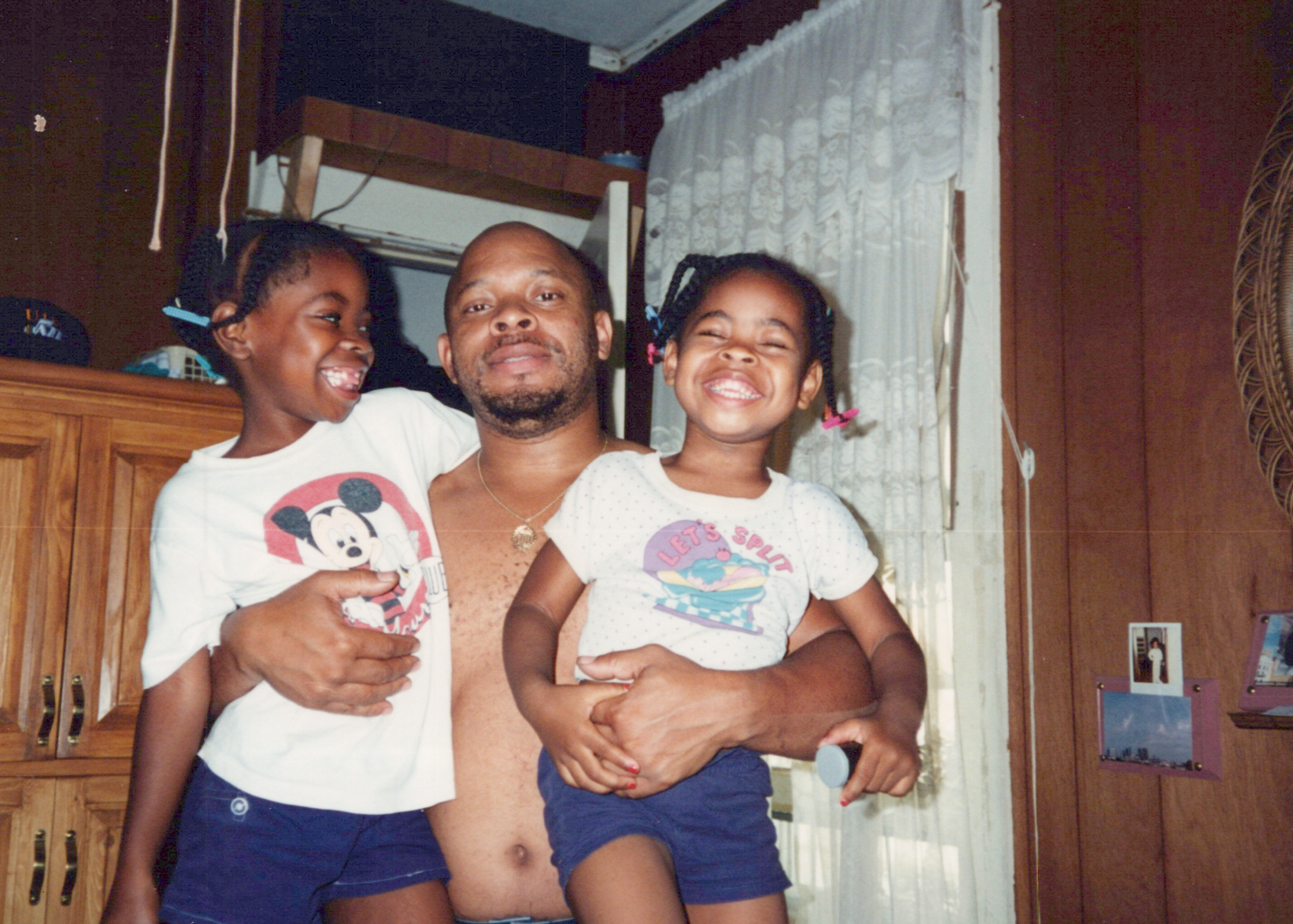 A man with dark skin tone inside a house holding his two smiling daughers.
