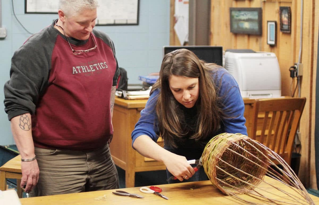 Two people with light skin standing near a table where one cuts an in-progress basket.