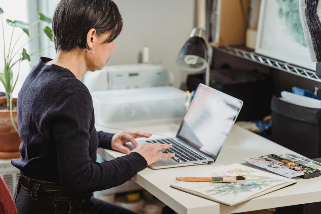 A person with medium skin tone and short dark hair sits at a desk typing on a laptop in a light-filled studio space, with watercolor paints and brushes nearby.