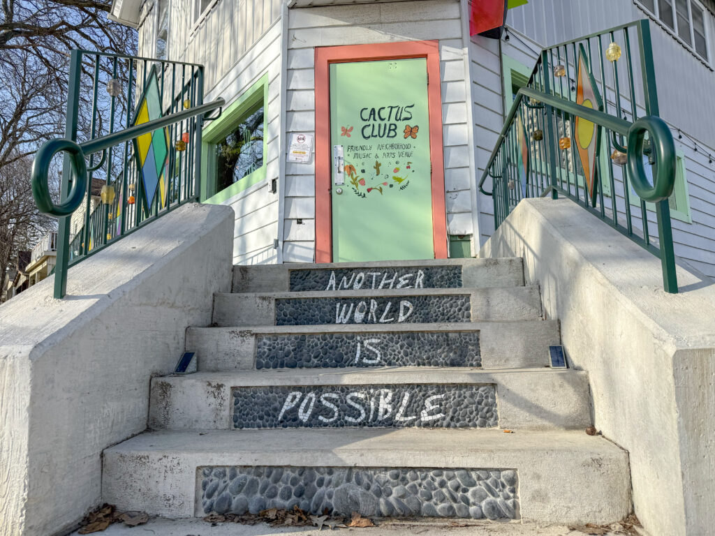 An outdoor concrete stair set with stone and tile mosaic. Each step has a mosaic word and together they read "another world is possible". The stairs lead up to a light green colored door that has hand-drawn signage that reads "cactus club" and "friendly neighborhood music and arts venue"