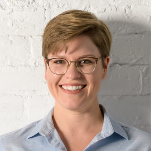 A headshot of a smiling person of light skin tone with short blonde hair wearing a blue blouse.