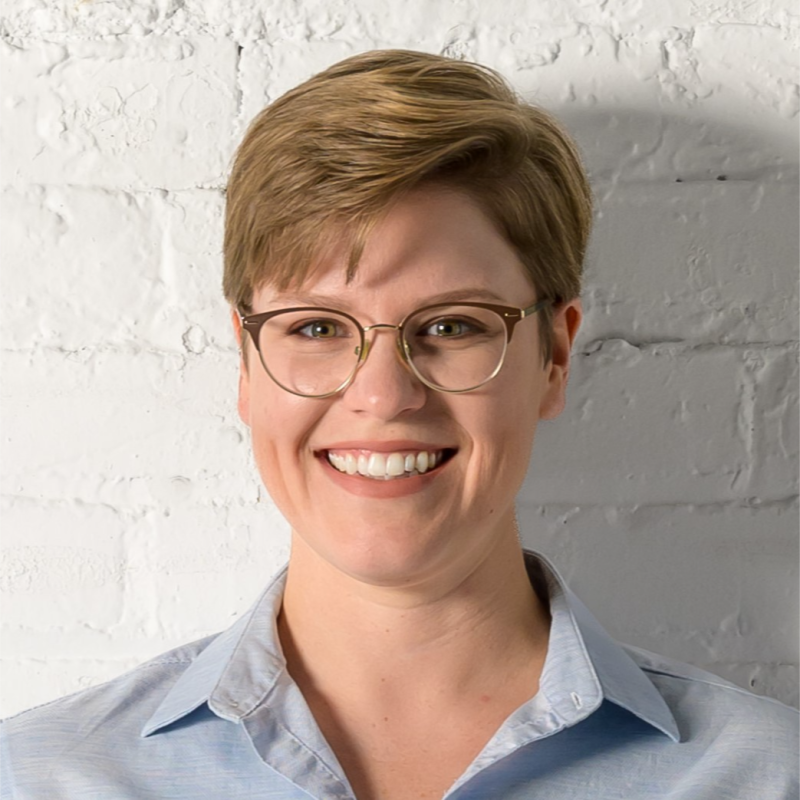 A headshot of a smiling person of light skin tone with short blonde hair wearing a blue blouse.