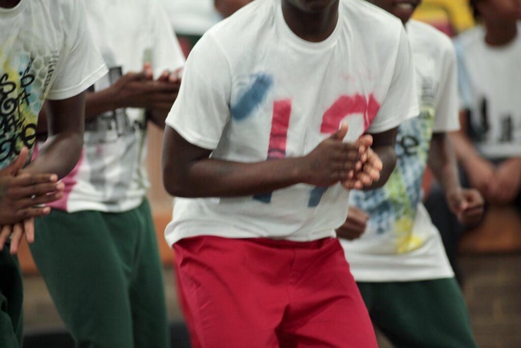 A row of young dancers caught mid-motion, framed from shoulders to knees. The dancer in the center wears a white t-shirt with a hand-painted red and blue design and red shorts, hands clasped in front of the chest. Others on either side wear similar white shirts with colorful graphic prints and green or red bottoms. The motion blur in the hands suggests a clap or rhythmic gesture.