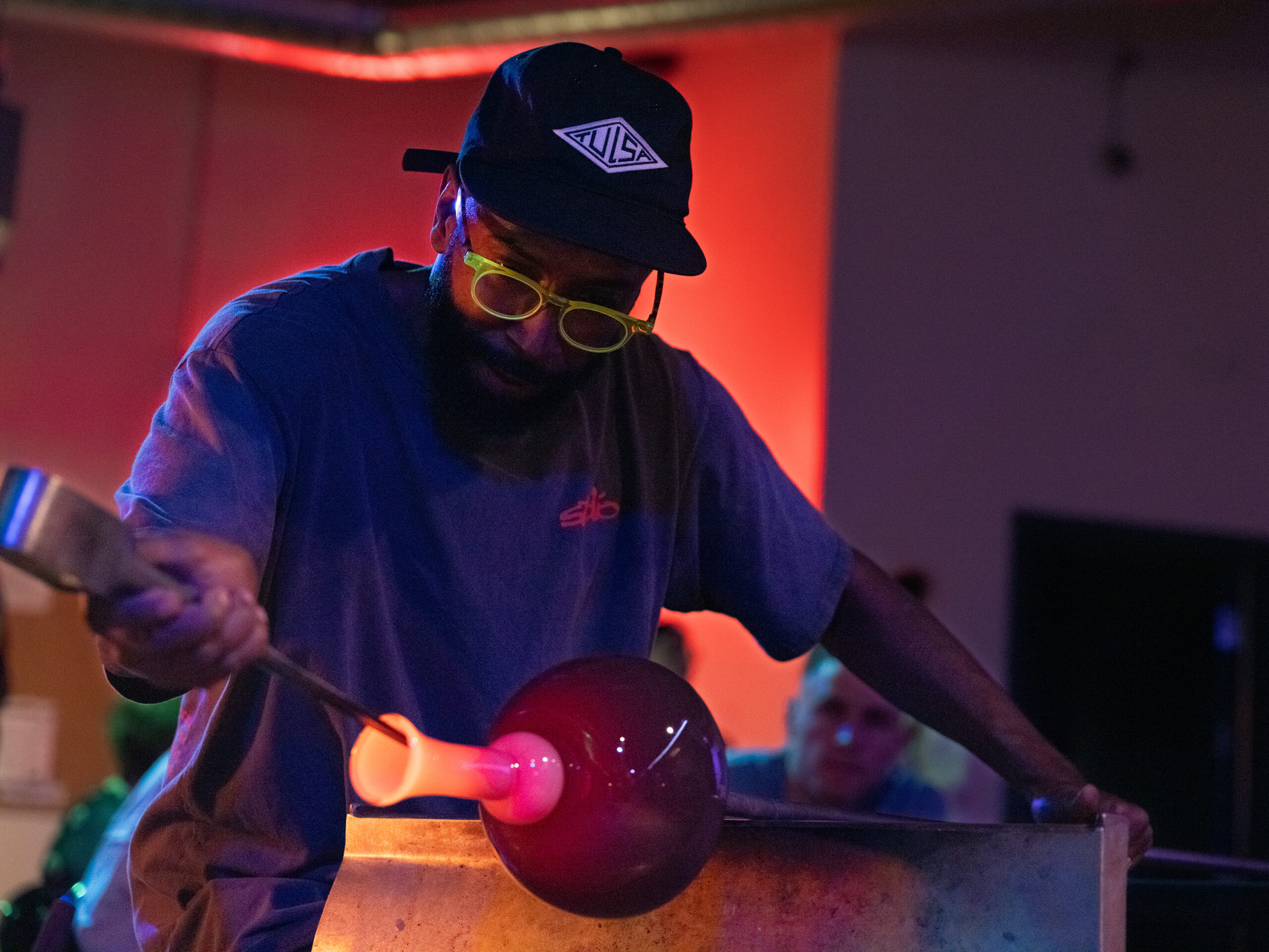 A person with dark skin wearing a flat him cap, t-shirt, and neon green glasses, shapes a piece of hot blown glass in dramatic lighting.