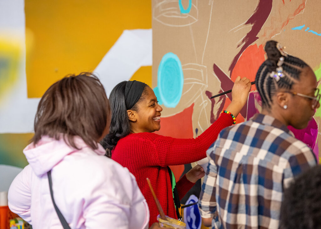 A person with medium-dark skin tone and black hair, wearing a red sweater, smiles joyfully while painting a large colorful mural. Two others paint alongside them.