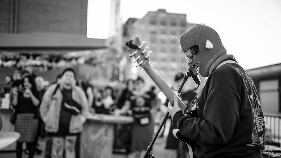 A person wearing a ski mask while singing and playing guitar for a crowd outdoors.