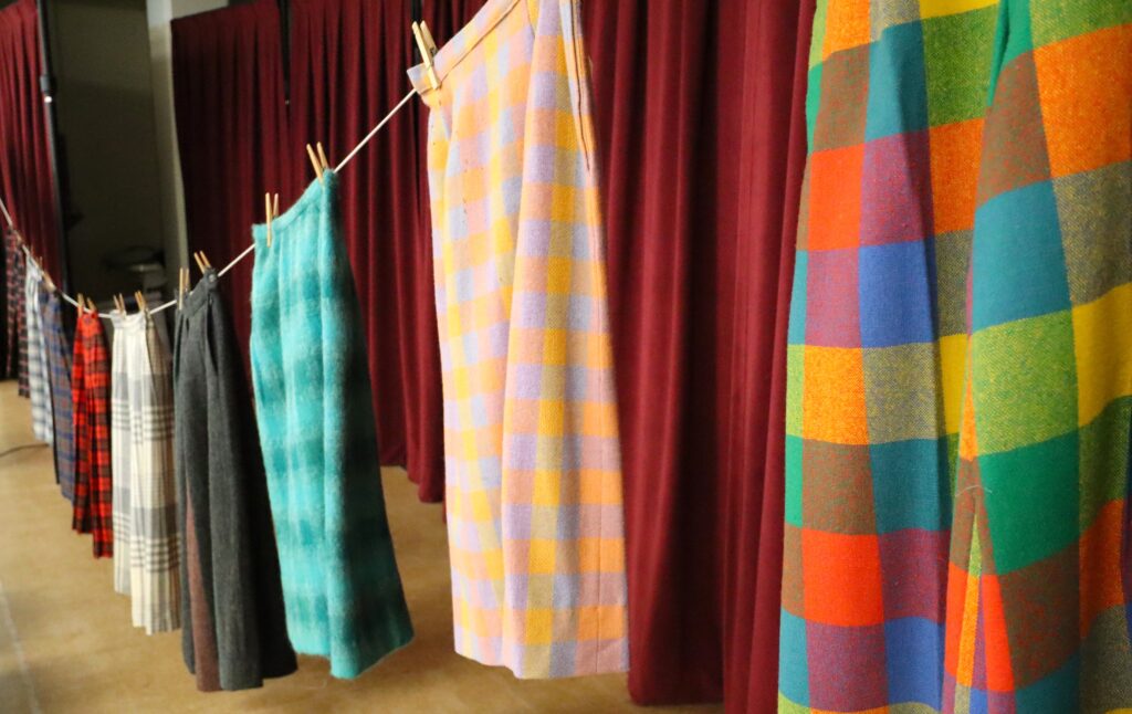 Several plaid skirts hanging on an indoor clothesline before a red curtain.