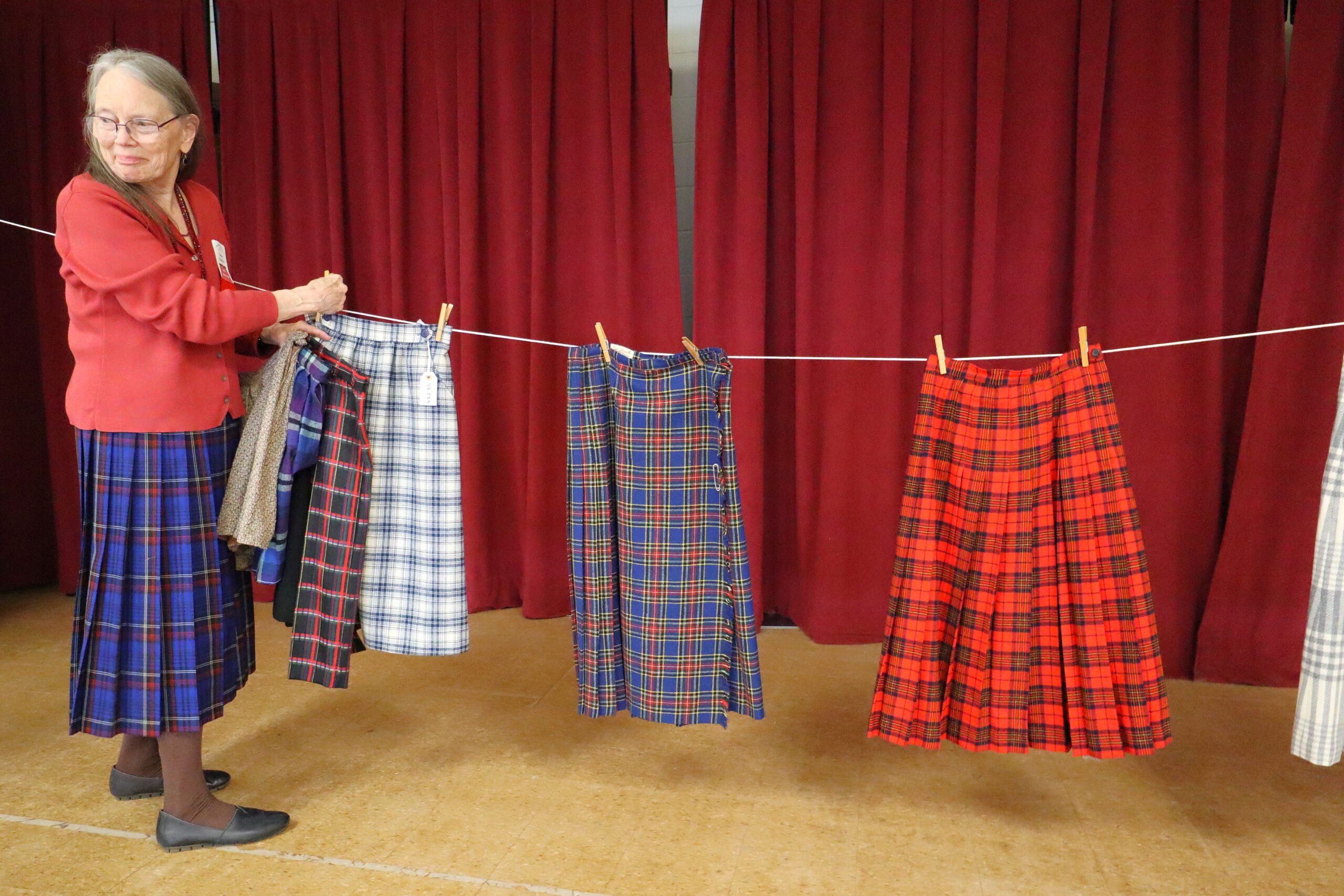 An older woman wearing a wool outfit hangs wool plaid skirts on an indoor clothesline.