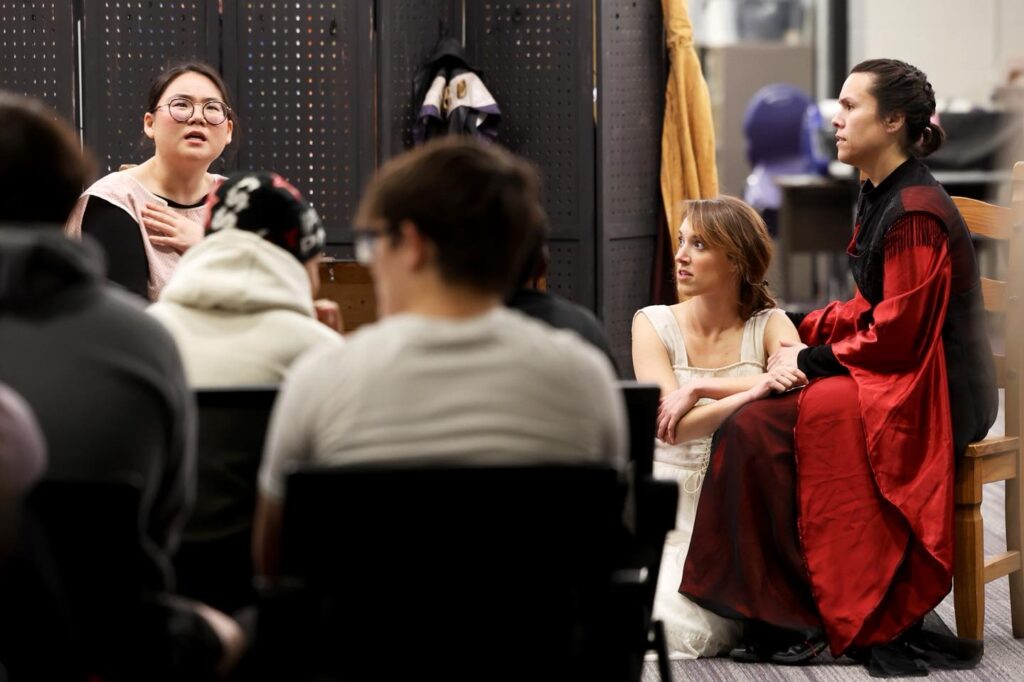 A woman with glasses and a pink top gestures with her hand to her chest, speaking with an upward gaze. Across from her, a young woman in a cream dress sits on the floor, leaning against the legs of another performer in a red and black gown seated on a wooden chair. Two figures in the foreground face away from the camera, watching. The setting looks like a rehearsal room with a dark perforated screen and costume pieces in the background.