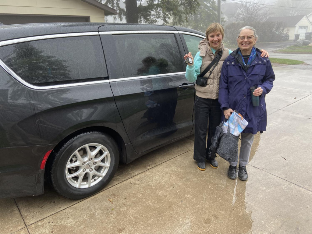 Two women standing and smiling next to a black minivan in a driveway.