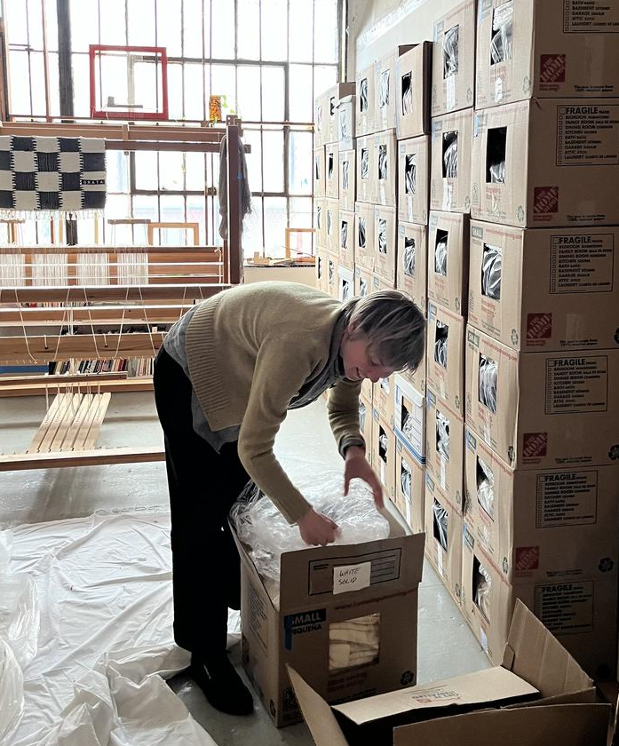 A woman with light hair and light skin reaches into a box amid a room full of boxes.