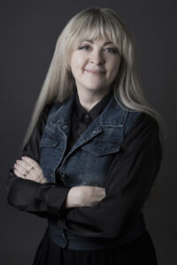 A person with light skin tone and long blonde hair with bangs smiles with arms crossed, wearing a black shirt layered under a denim vest.