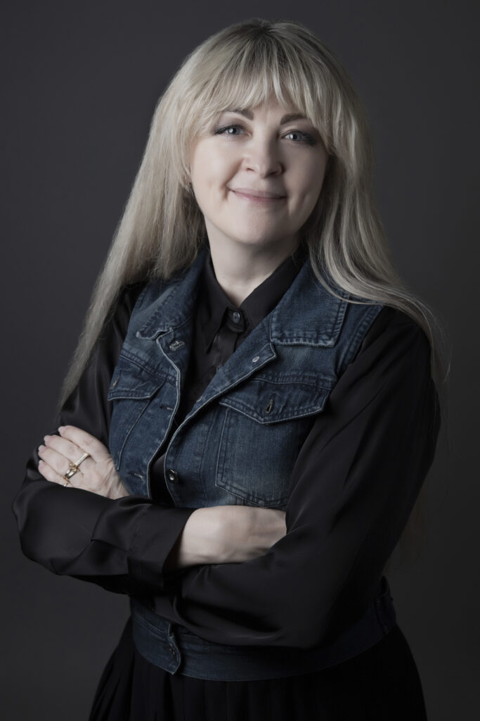 A person with light skin tone and long blonde hair with bangs smiles with arms crossed, wearing a black shirt layered under a denim vest.