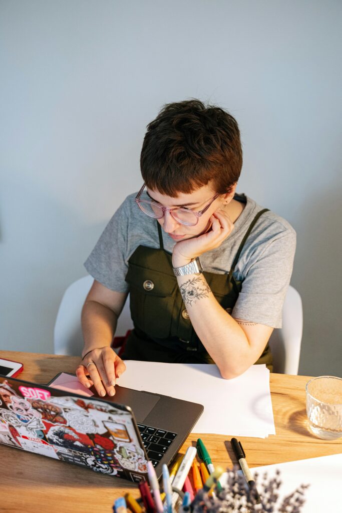 A person with light skin tone and short brown hair wearing a gray t-shirt and green overalls rests their head on their arm while using a laptop covered in stickers.