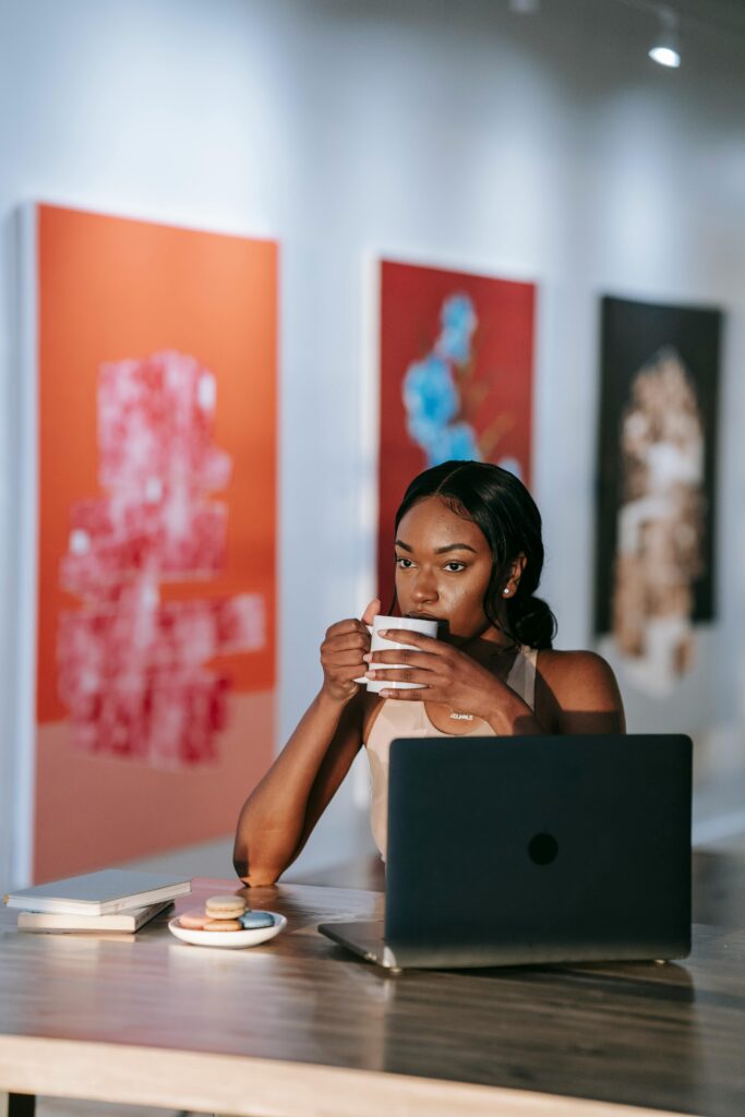 A person with dark skin tone and long, wavy black hair sits at a laptop drinking a cup of coffee introspectively.