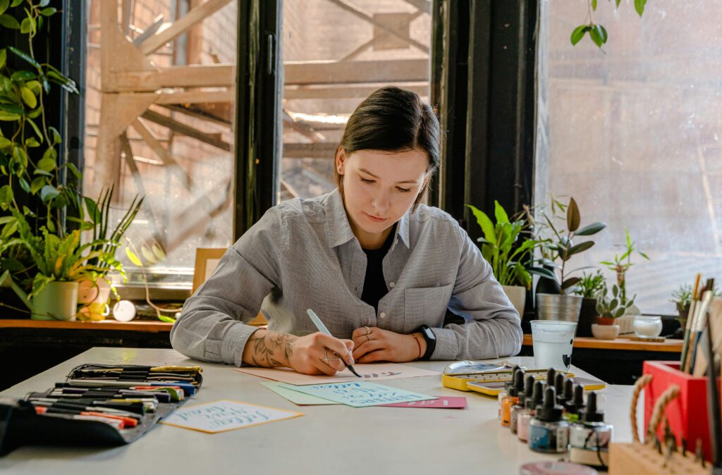 A person with light skin and dark hair pulled back practices calligraphy at a well-lit desk surrounded by plants and art supplies.