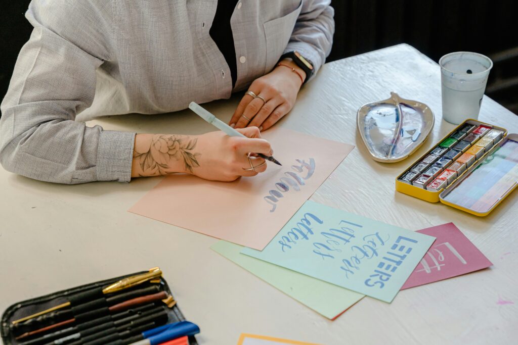 A person with light skin tone with a black flower tattoo wearing a gray blouse practices watercolor calligraphy at a well-lit desk.