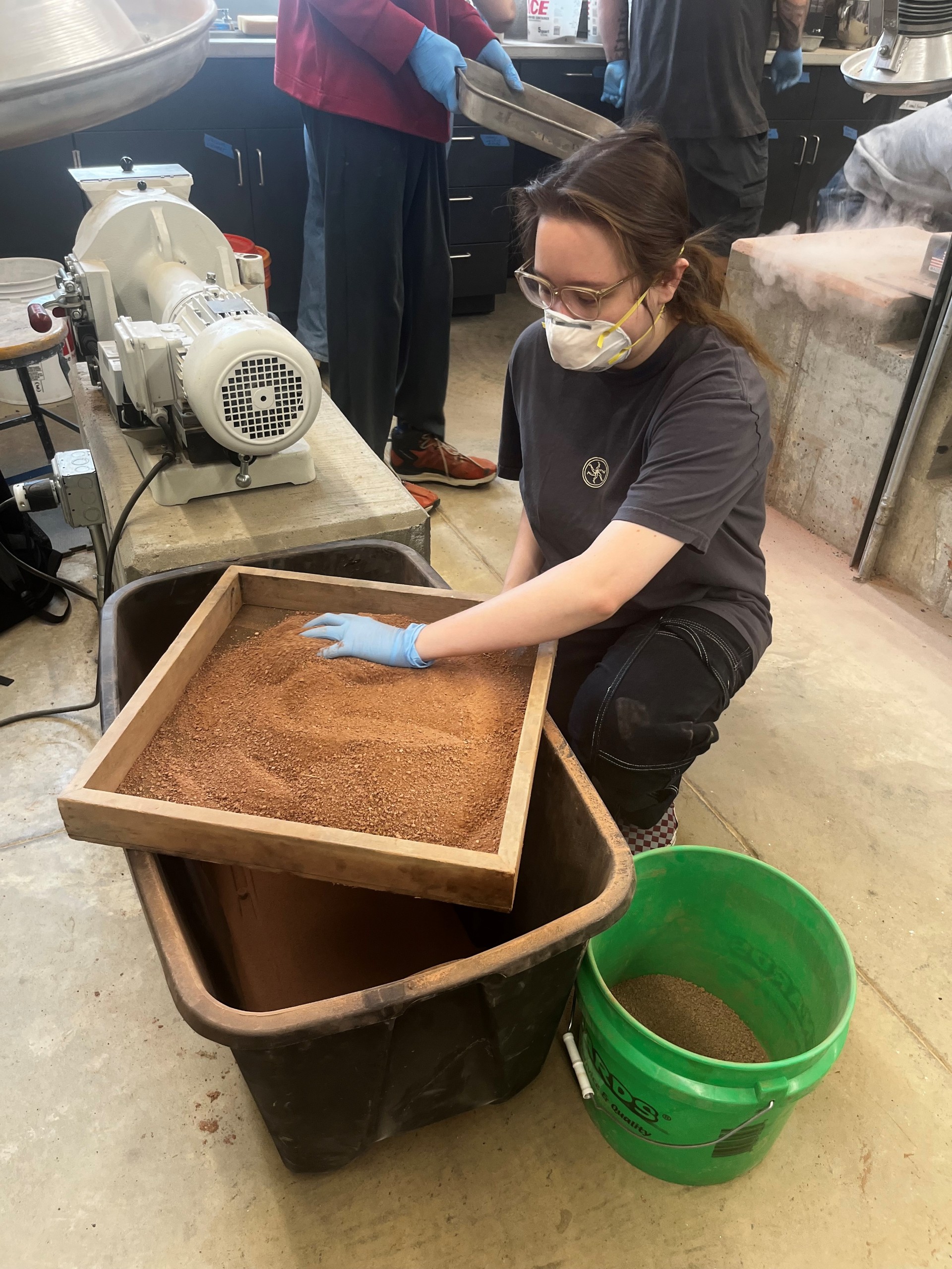 A person wearing a mask and processing clay indoors with large buckets.