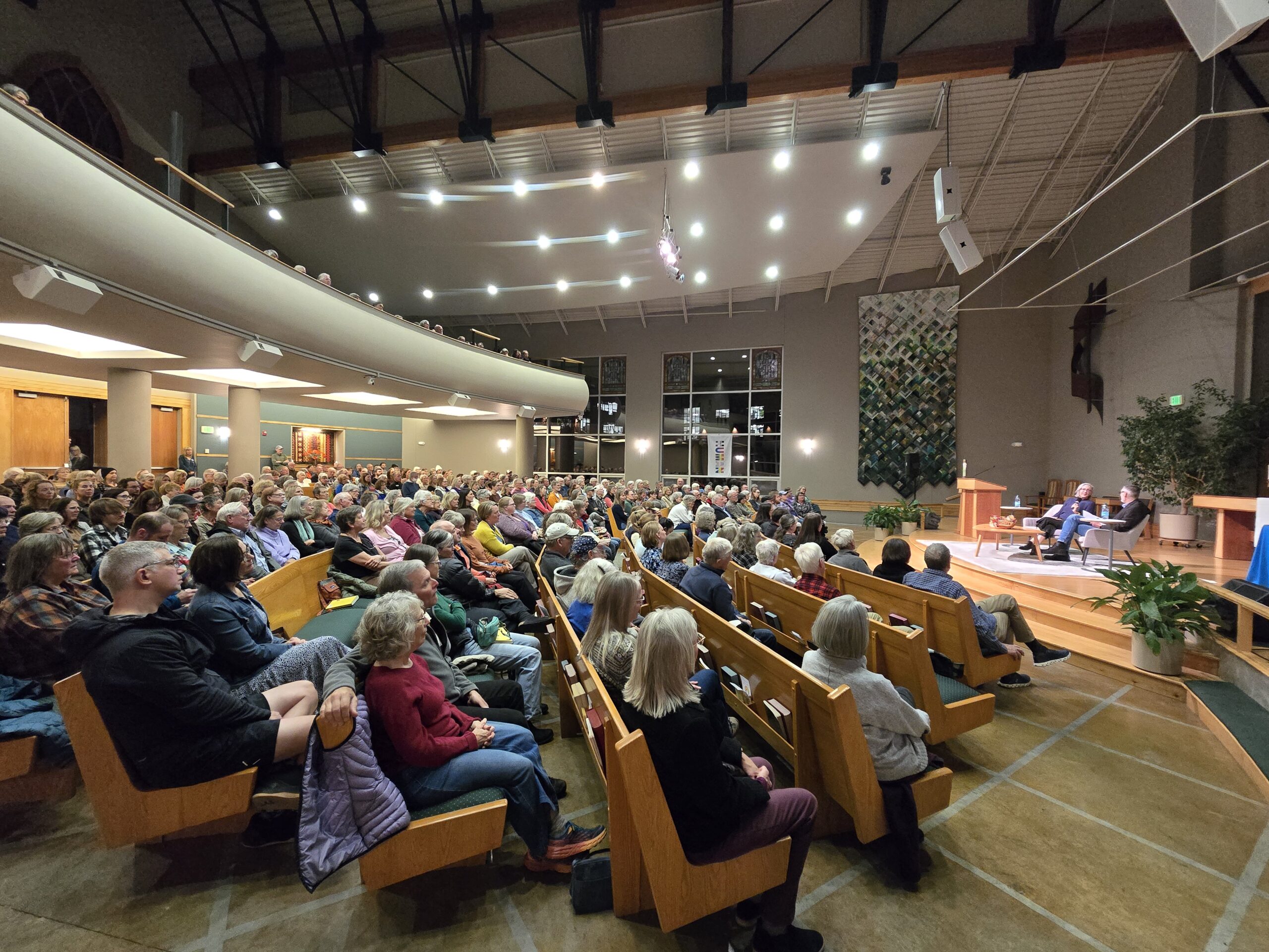 A full room of audience seated on pew benches looks on at the stage where two people are seated and in conversation.