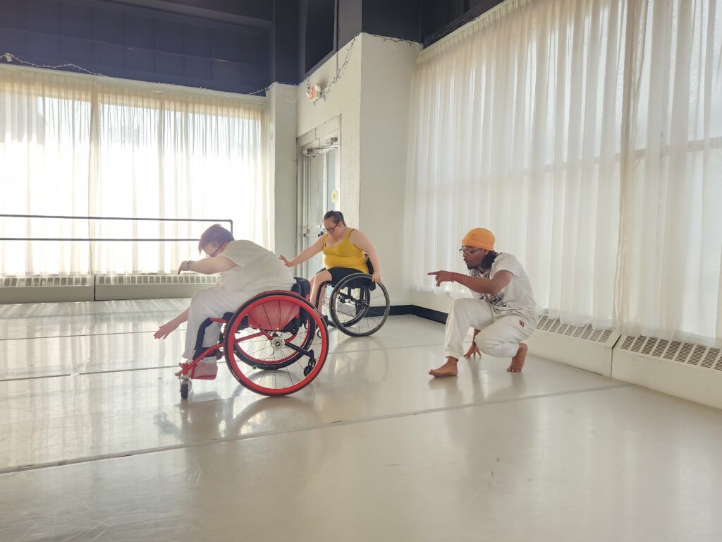 Three dancers rehearse in a bright white room. Two of the dancers use a wheelchair.