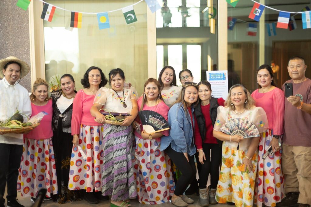 A large group of people with medium skin tones poses together beneath international flags at a Festival of Cultures event, several holding decorative fans and produce.