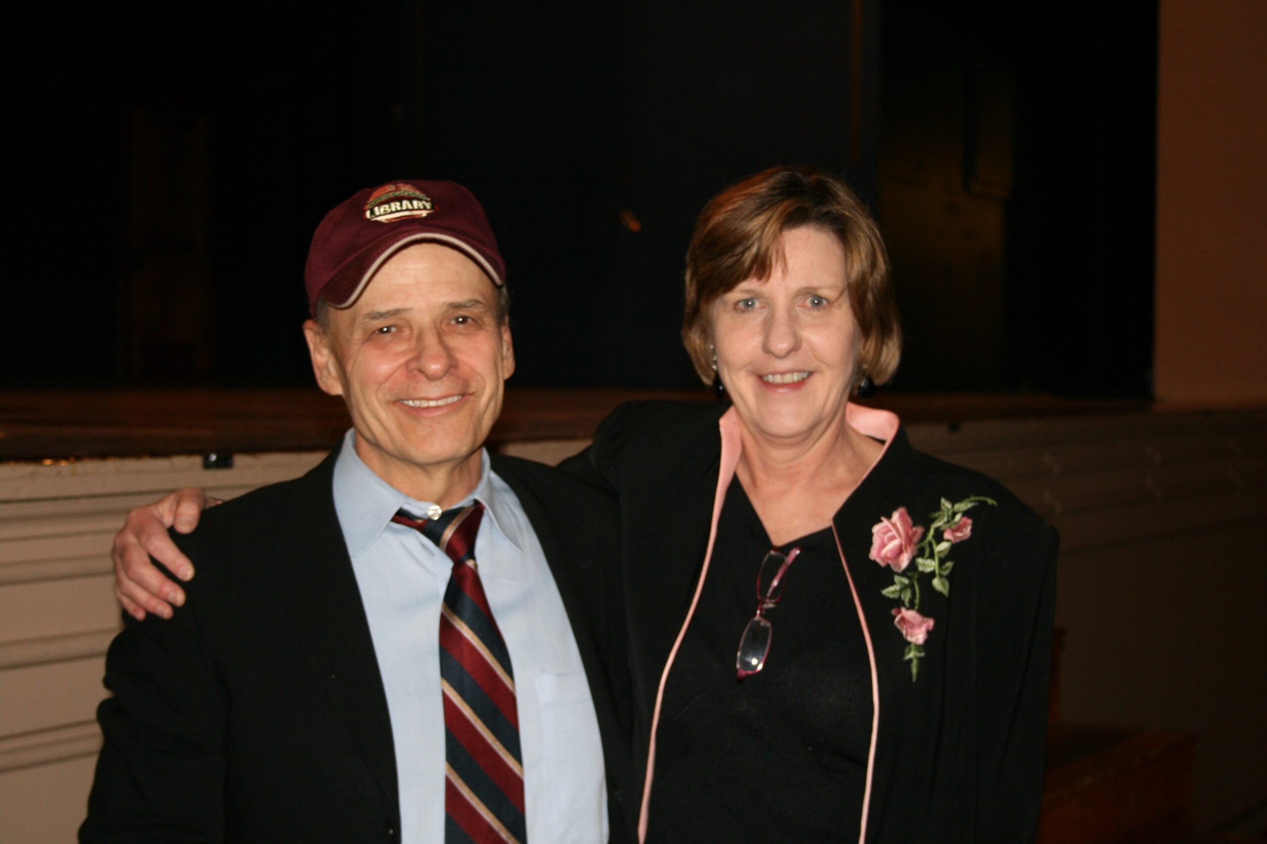 Two people standing next to each other and smiling. One is wearing a suit and tie with a baseball cap and the other is wearing a dark top with a dark jacket with a floral embroidery.