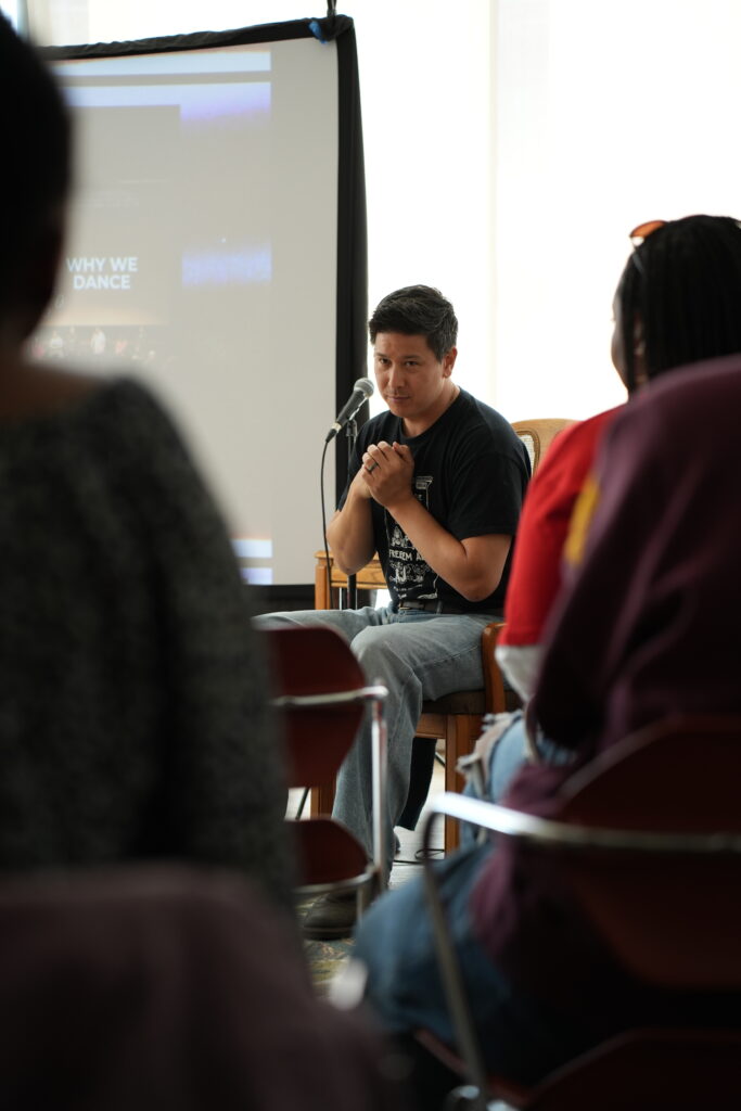A person with black hair and medium skin tone wears a black t-shirt and a wedding band. The person hold their hands together at their chest and looks solemnly at a crowd in front of a project screen next to a microphone.