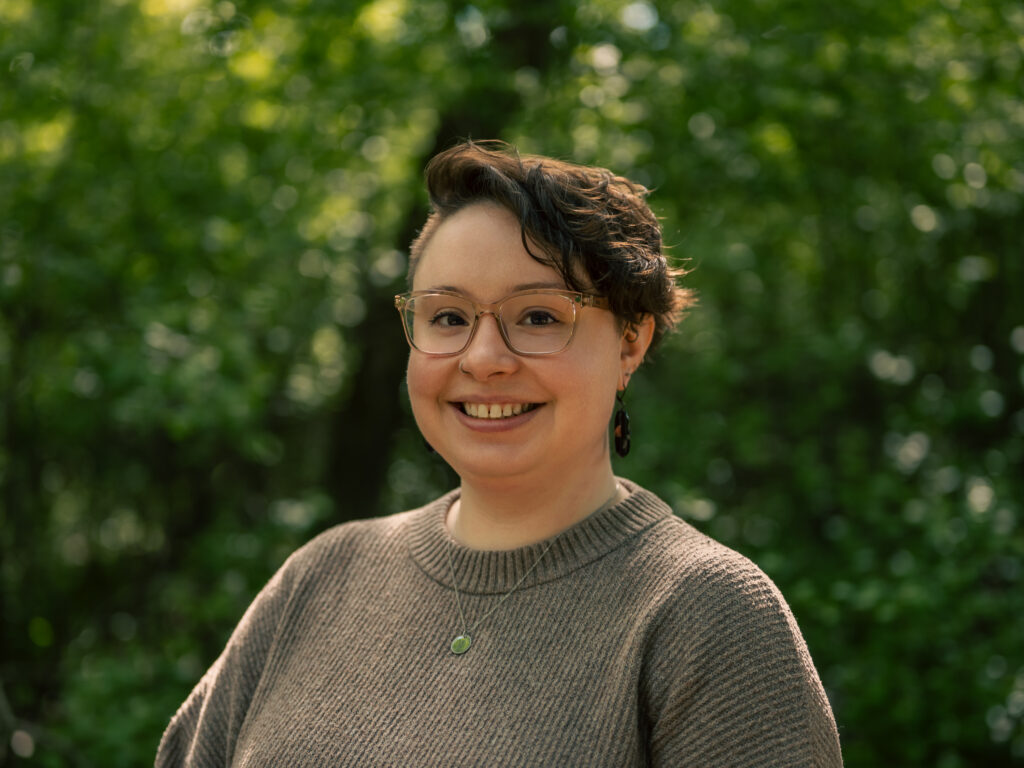 Headshot of a person with short, curly dark brown hair with auburn highlights, wearing pink-framed glasses, a taupe ribbed sweater, a small green pendant necklace, and dark drop earrings. They smile warmly against a blurred green leafy background.