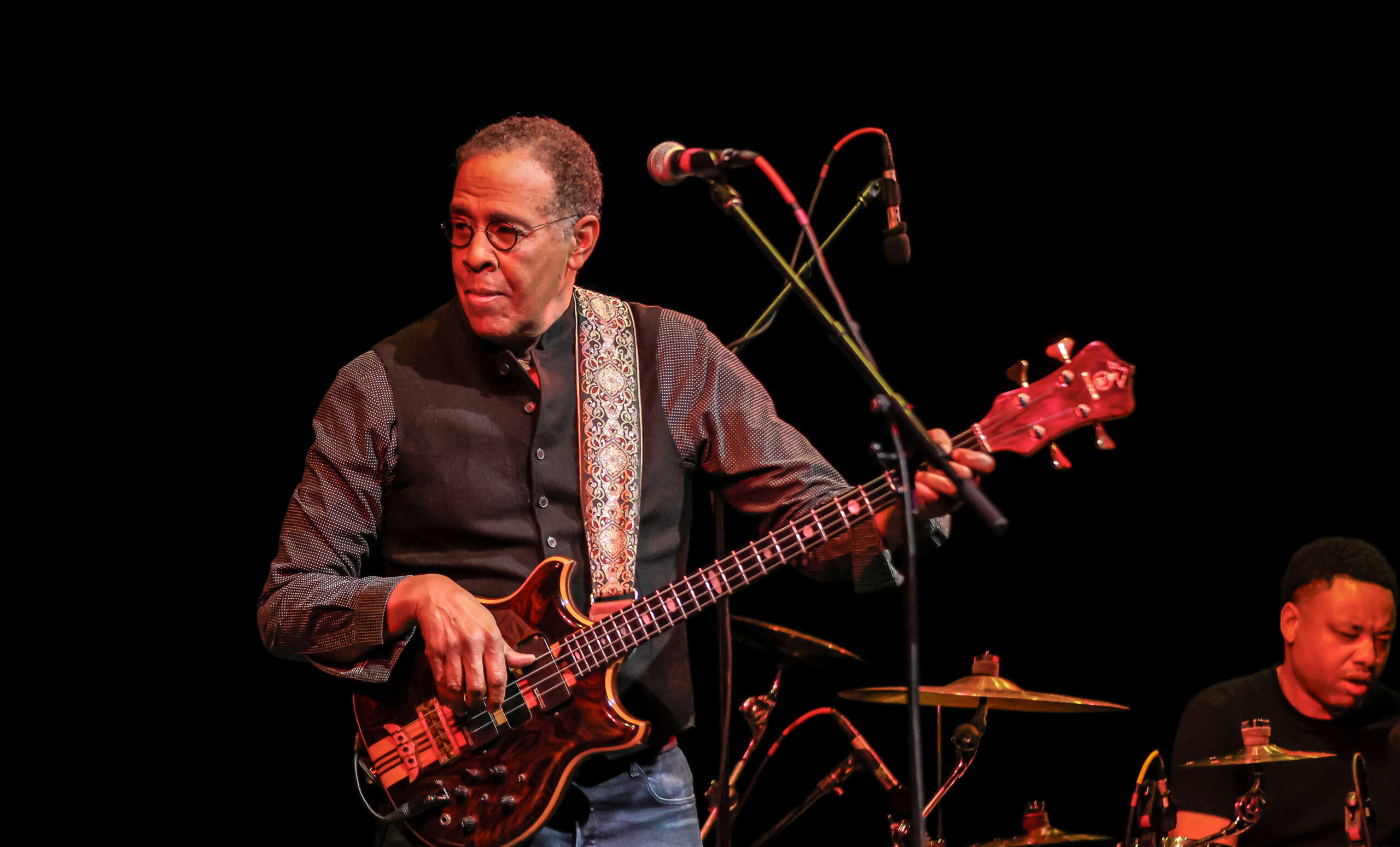 An older adult with dark skin tone and glasses plays an ornate bass guitar on a darkened stage; a drummer is visible in the background.