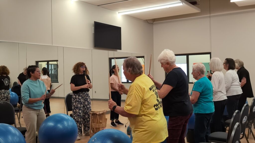 A group of older adults and instructors stand in a mirrored exercise room, each holding drumsticks and drumming on large blue exercise balls.