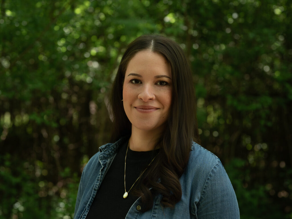 Headshot of a person with long, straight dark brown hair, wearing a denim jacket over a black shirt and a small gold pendant necklace. They smile gently against a blurred green tree background.