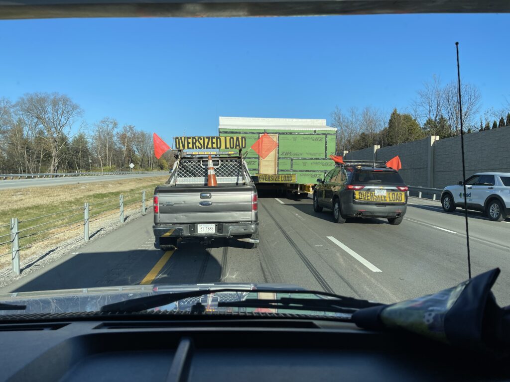 Cars with signs that read "oversized load" on a highway follow a semi trailer hauling a house structure