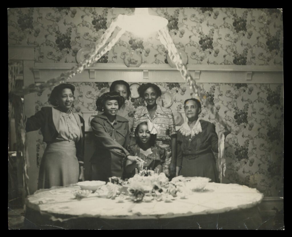 A black and white archival photo of a child's birthday with cake and candles on the table in from of them and five adults standing around