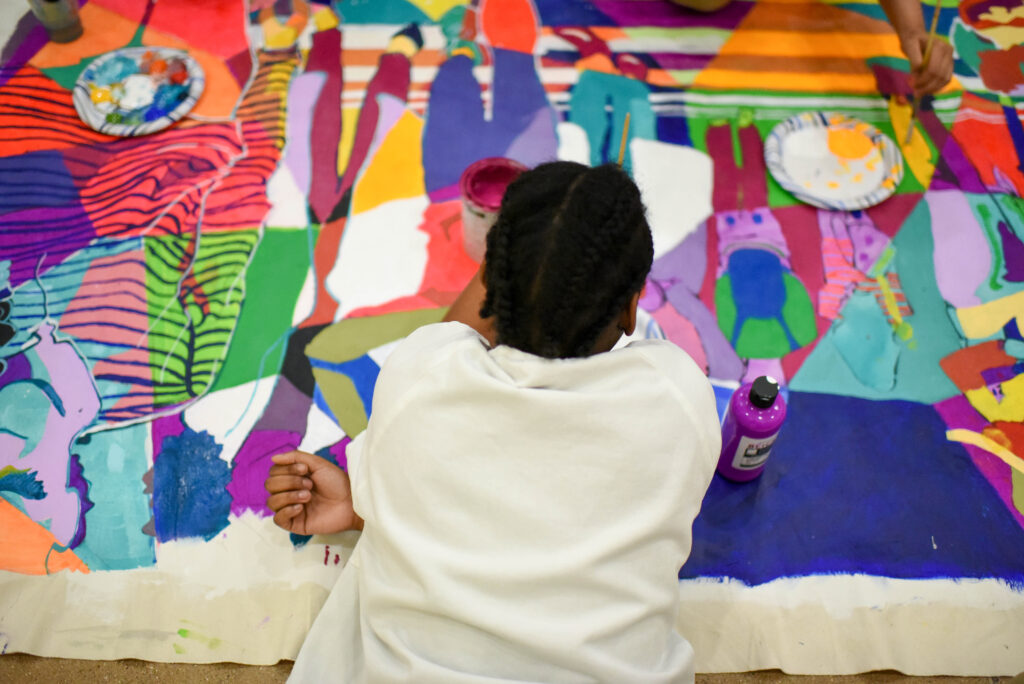 A person kneeling to paint a large, brightly colored mural laid out across the floor.