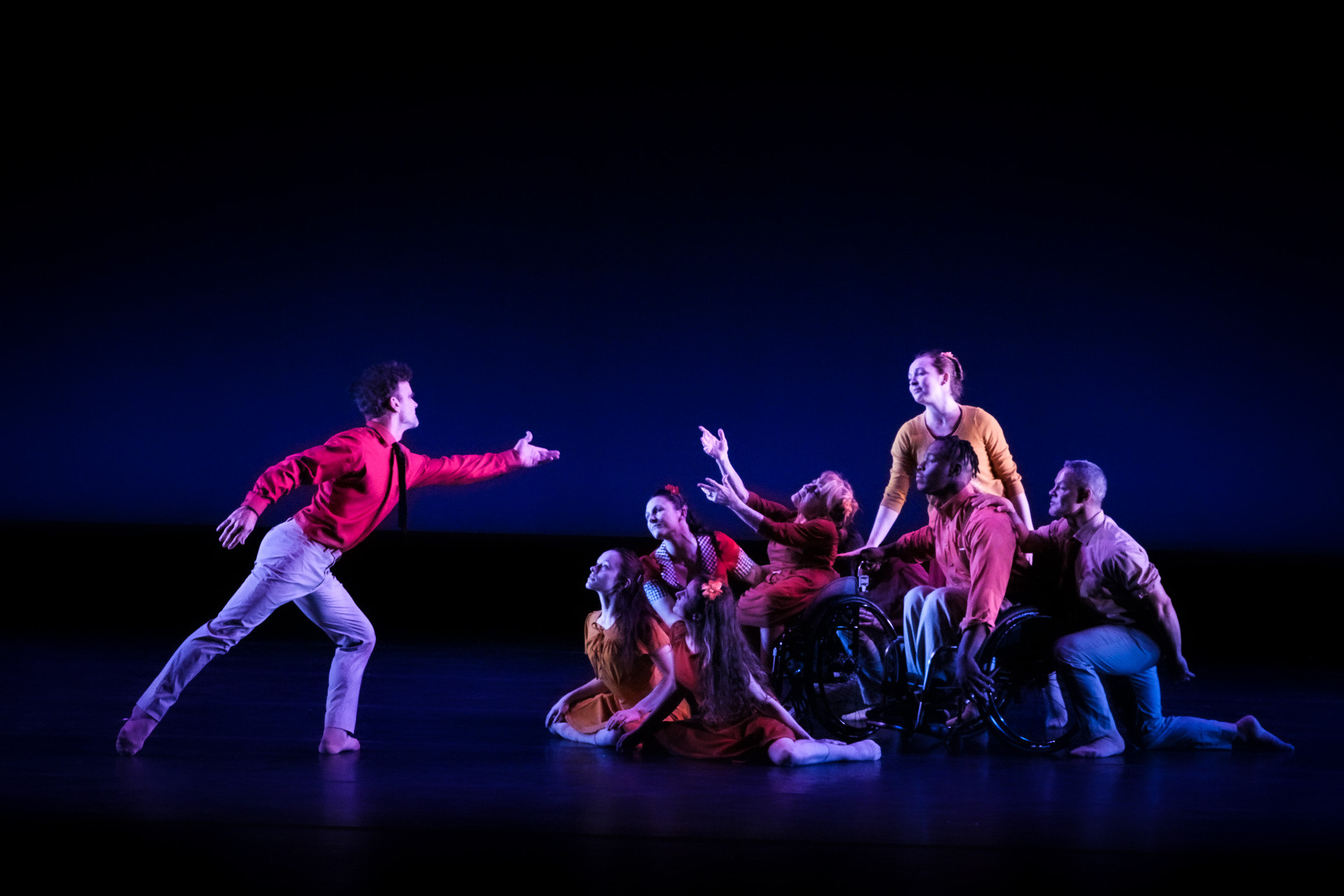 An ensemble of dancers poses with company dancer Mary Verdi-Fletcher reaching toward solo dancer Brian Murphy who is lunging and reaching toward her.