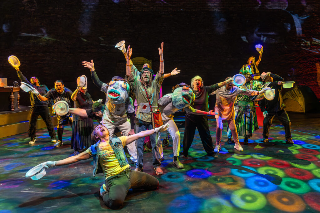 A large ensemble cast of diverse performers in eclectic costumes—including two large puppet heads—throw their arms wide in a climactic moment on a colorfully lit stage.